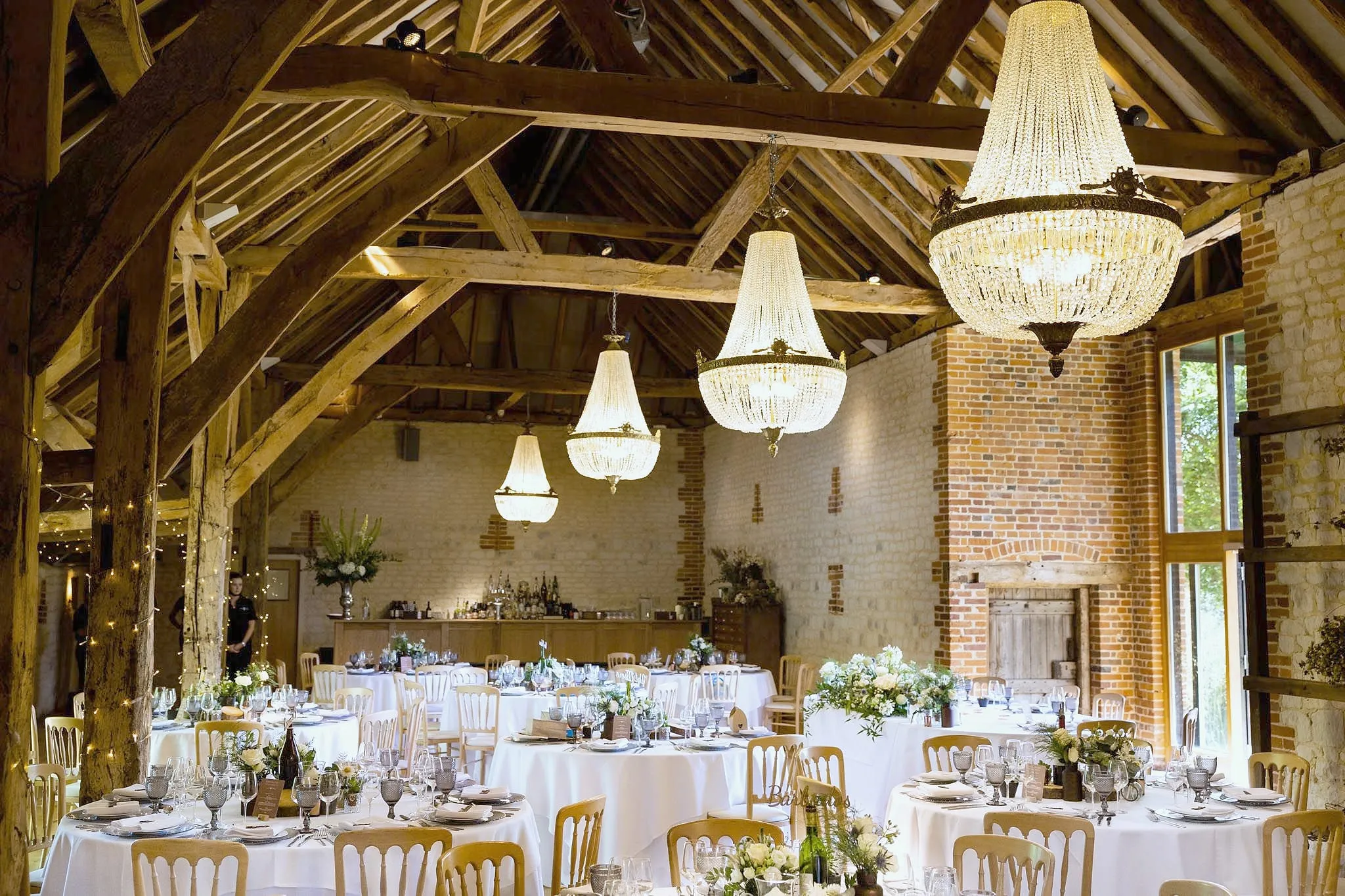 The barn laid for dinner with round tables, white linen and crystal chandeliers under oak beams