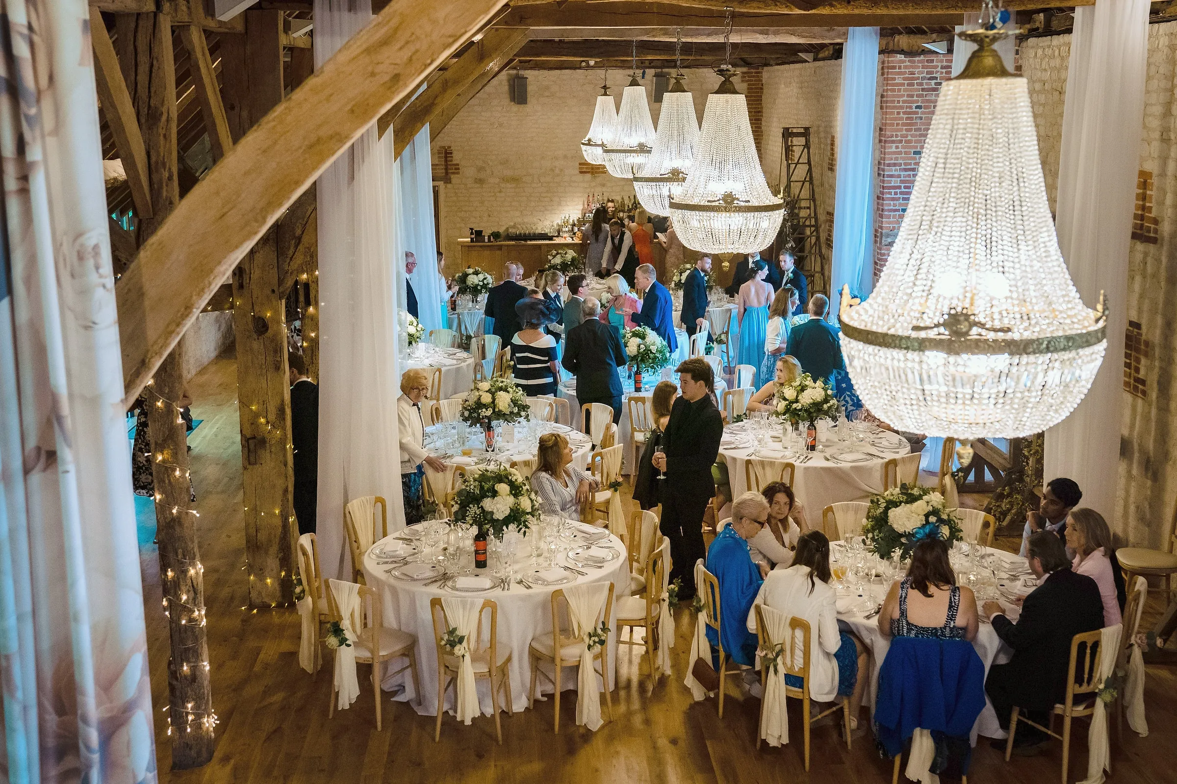 Guests finding their seats at round tables in the barn beneath large crystal chandeliers