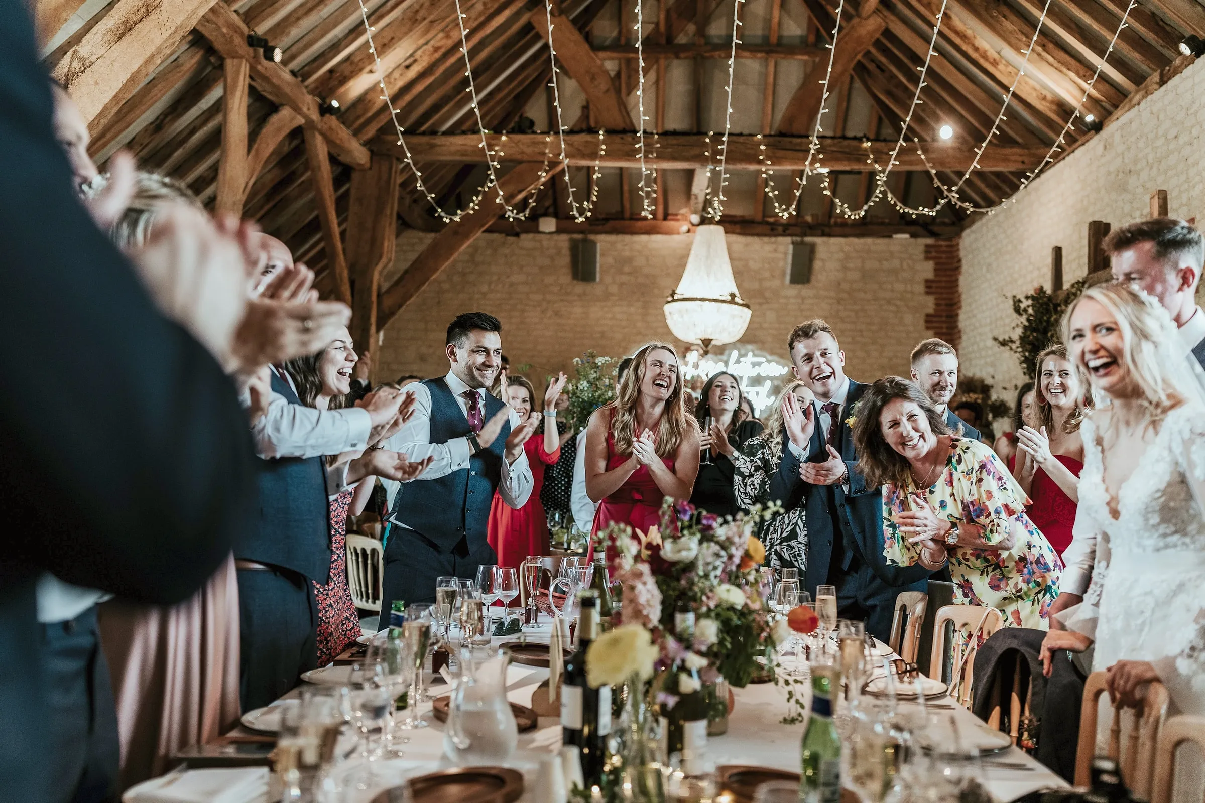 Guests at long tables in the barn raising their glasses and cheering under the fairy lights