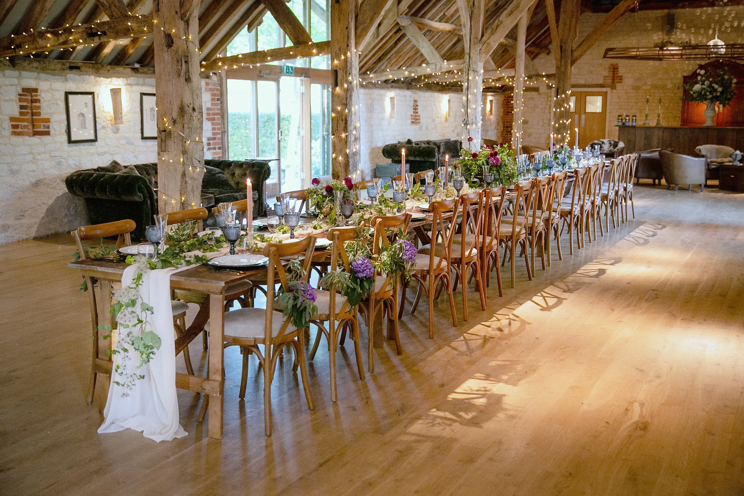 A single long banquet table running the length of the barn with candles, greenery and evening sun on the floor