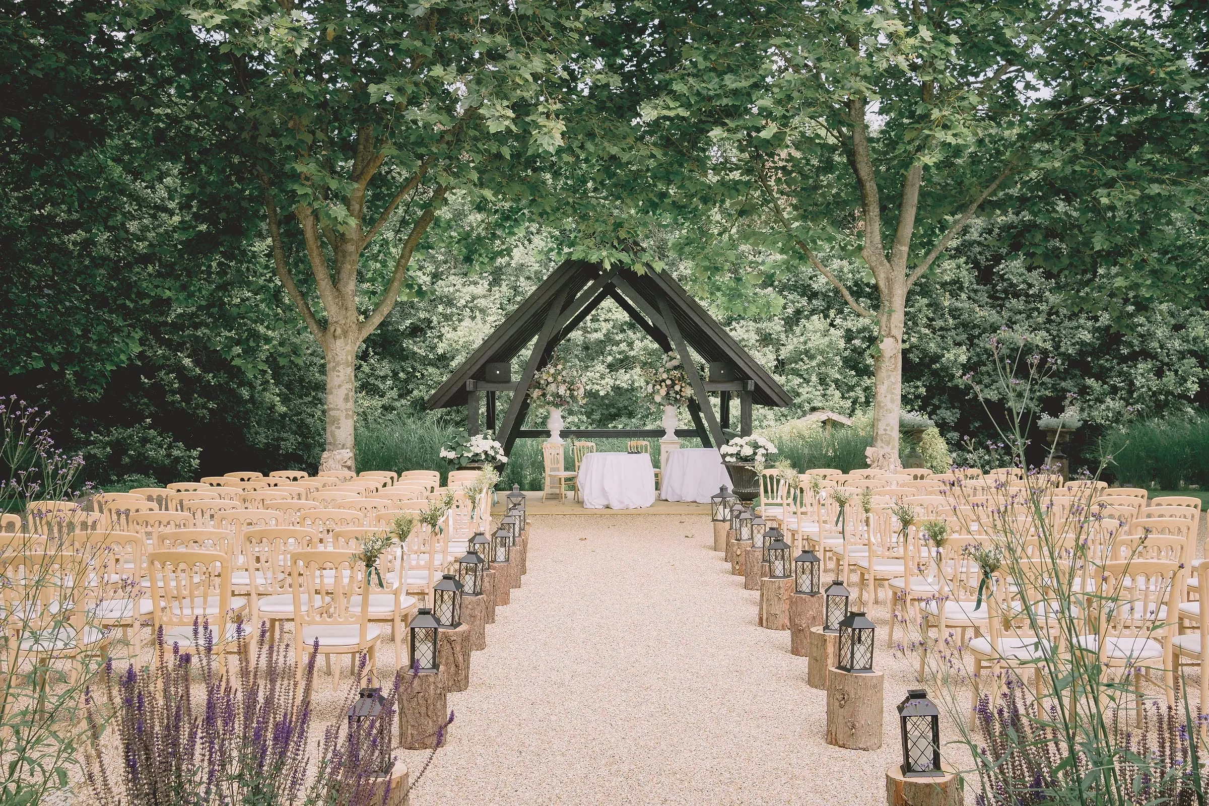 Rows of wooden chairs and lanterns along a gravel aisle to the oak ceremony shelter beneath plane trees