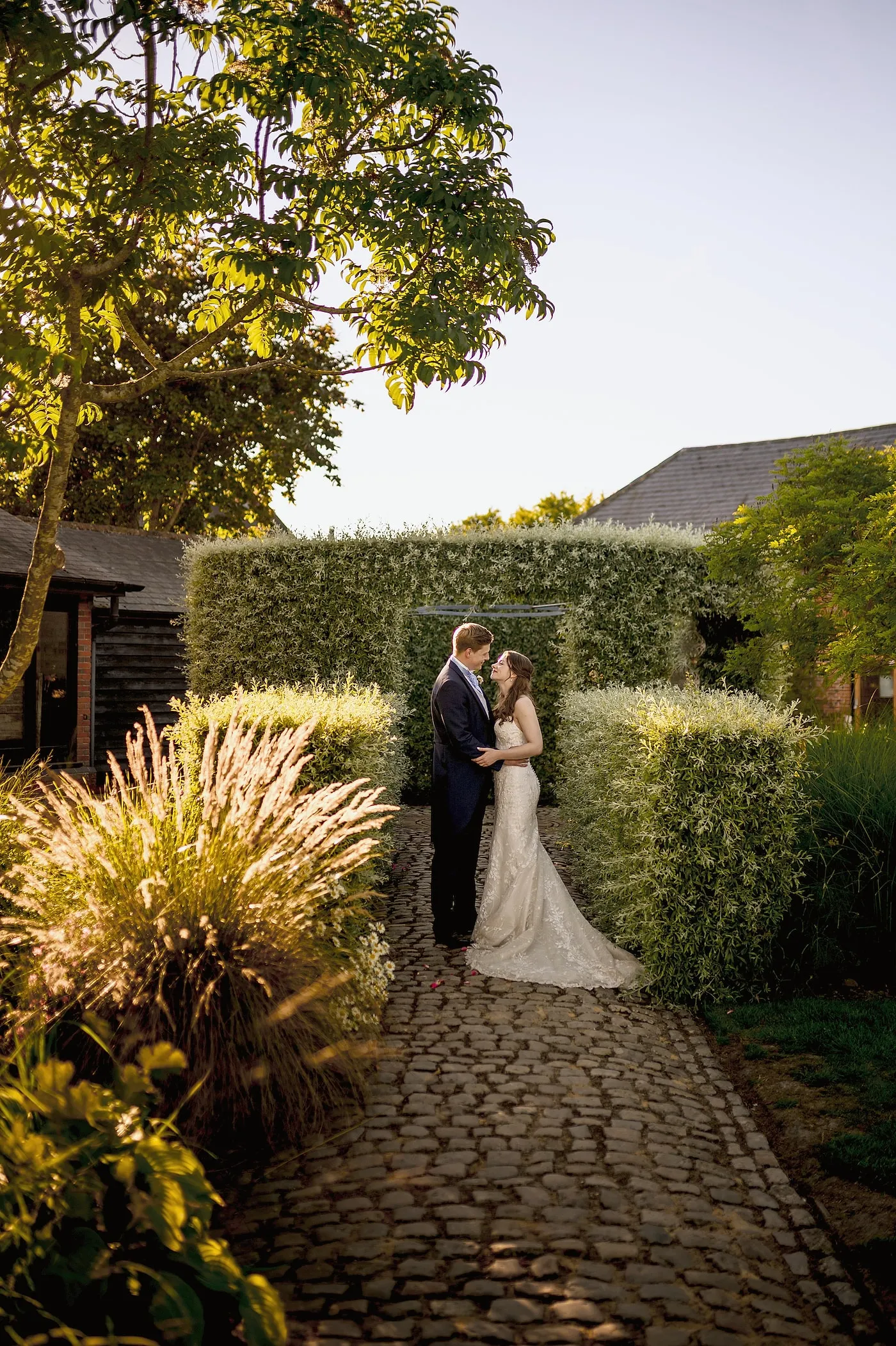A bride and groom kissing on a cobbled path between clipped hedges in evening light