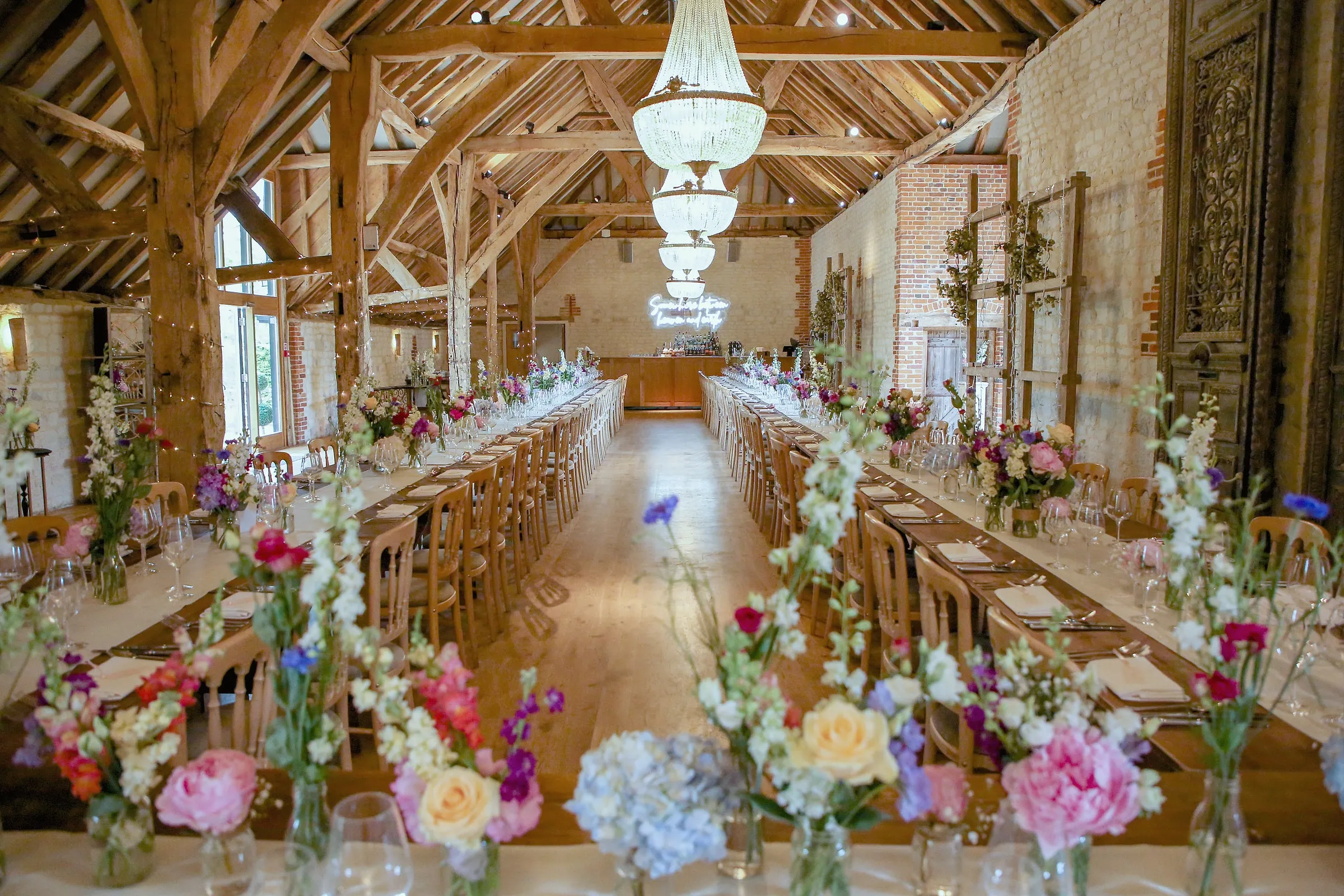 Long tables in the barn dressed with jugs of summer flowers beneath crystal chandeliers and oak trusses