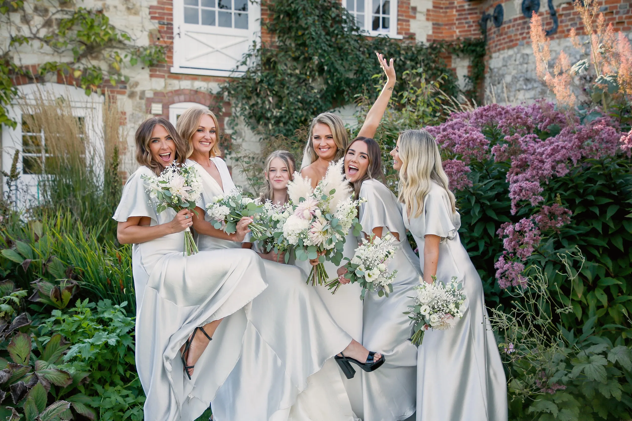 Bride and bridesmaids in pale satin laughing with bouquets in front of the flint house and late summer flowers