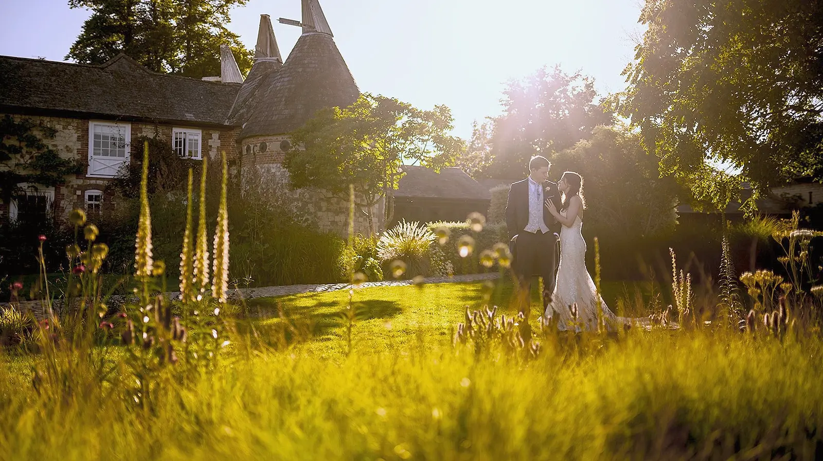 A couple in long grasses at golden hour, with the oast houses and trees behind