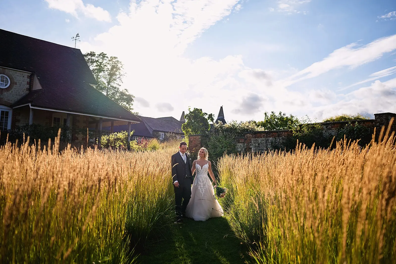 Couple walking between tall golden grasses in evening sun, the oast house roof behind them